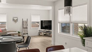 A modern living room with a flat-screen TV, a dining area with a round table and chairs, and large windows with blinds.