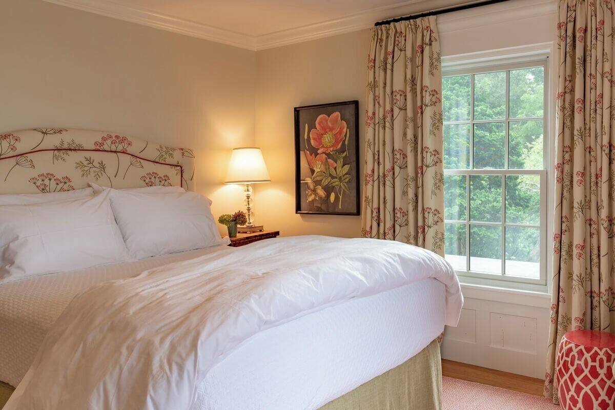 A neatly made bed with white linens, a bedside lamp, a floral painting on the wall, and a window with curtains.