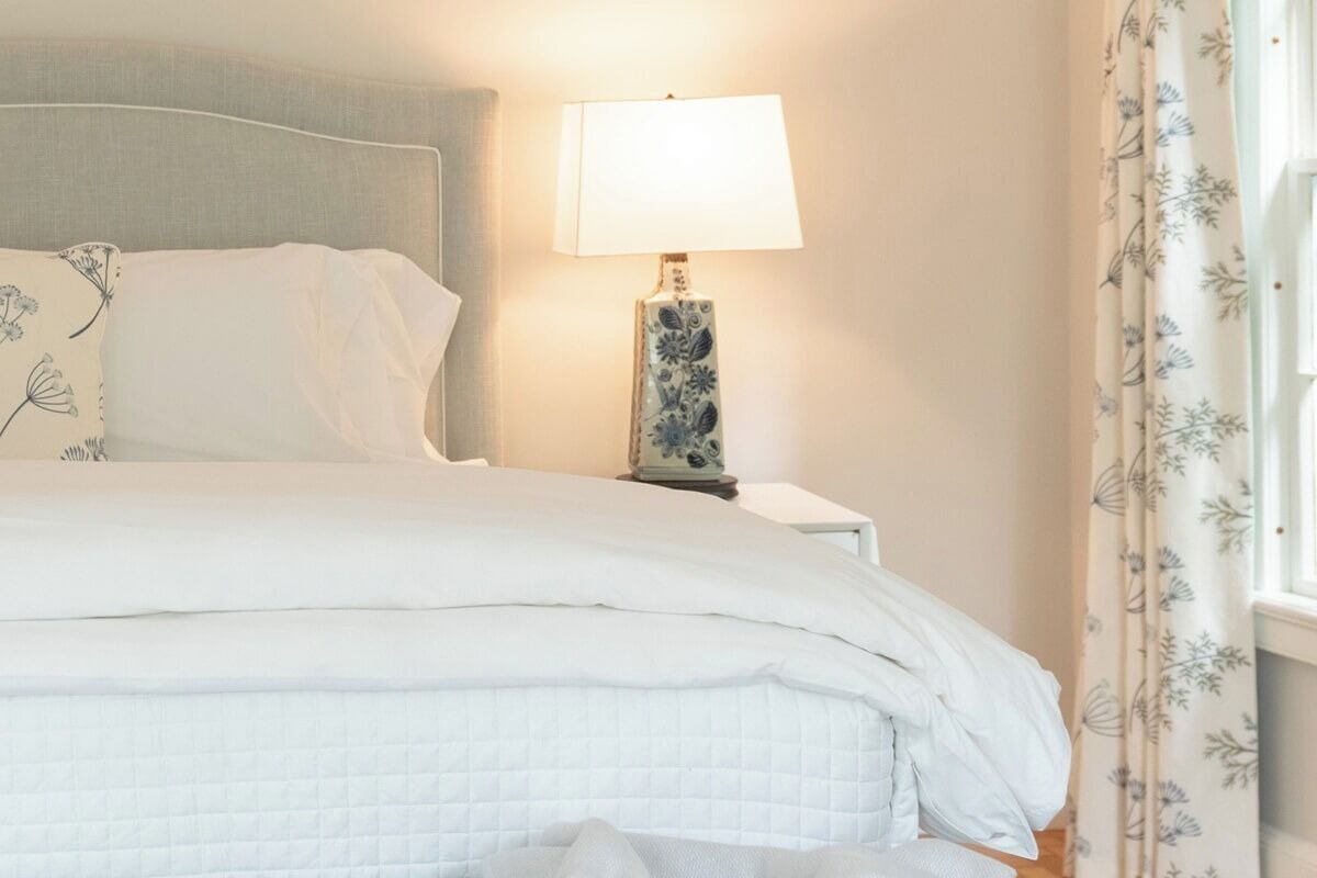 Una cama bien hecha con ropa de cama blanca y una lámpara con estampado floral sobre una mesita de noche.