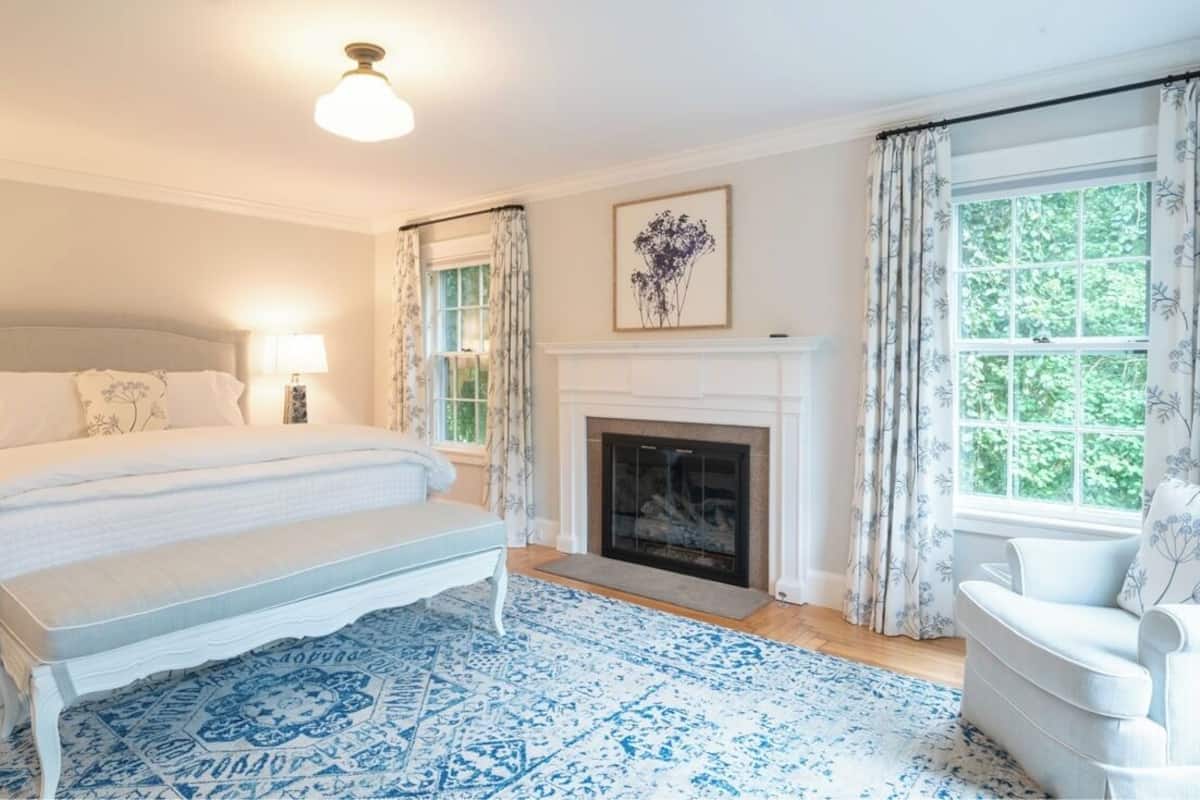 A bedroom with a large bed, a bench, a fireplace, and a window with curtains.