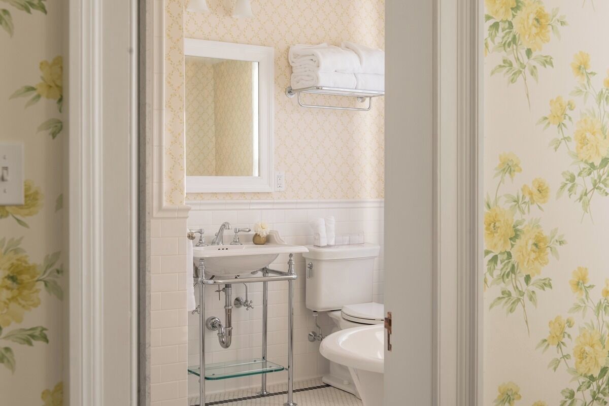 A bathroom with a white sink, mirror, and toilet. There is a towel rack above the toilet and a glass shelf below the sink.