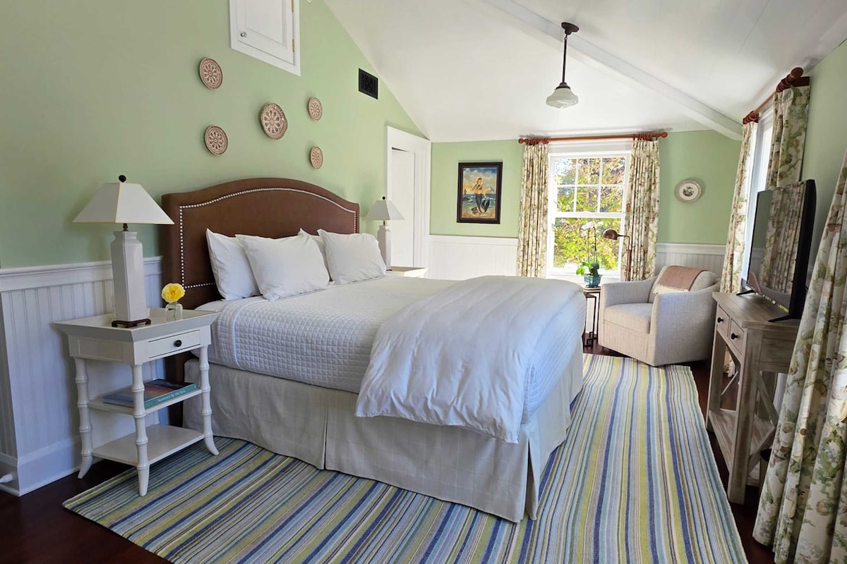 A bedroom with a large bed, a bedside table, a chair, a desk, a window with curtains, and a painting on the wall.