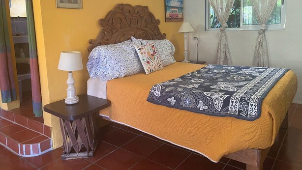 A bedroom with a wooden headboard, a bed with a floral bedspread, a bedside table with a lamp, and a window with curtains.