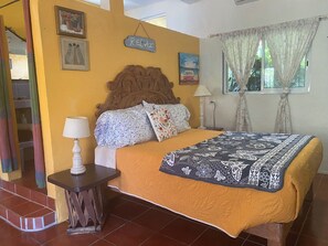 Un dormitorio con cabecera de madera, cama con cubrecama floral, mesita de noche con lámpara y ventana con cortinas.