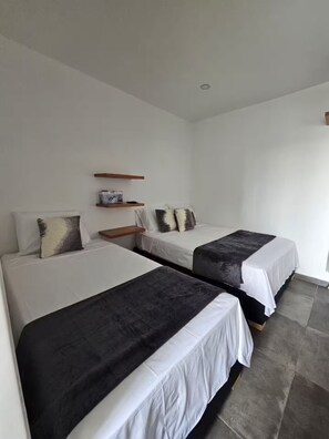 Habitación de hotel con dos camas, ropa de cama blanca y una pequeña estantería con un libro.