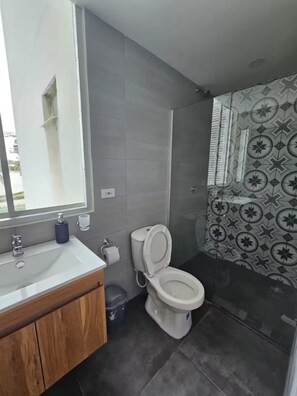 Un baño moderno con inodoro, lavabo y espejo. Presenta baldosas grises, un revestimiento de ducha estampado y un mueble de tocador de madera.