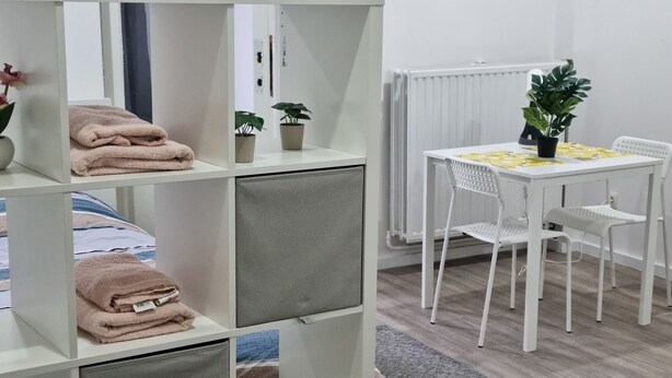 Une chambre d’hôtel moderne avec une armoire blanche, un coin repas comprenant une table et des chaises blanches, et un tapis gris sur le sol.