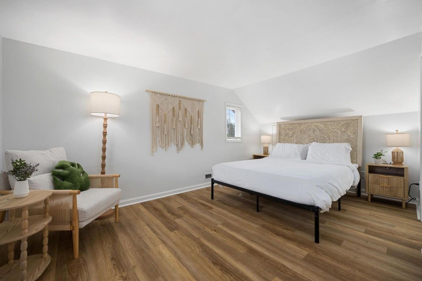 A bedroom with a bed, bedside tables, a lamp, a macrame wall hanging, and a green plant.