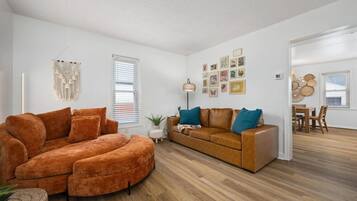Un salon spacieux avec un grand canapé orange, un fauteuil orange plus petit, un canapé marron agrémenté de coussins turquoise, et un coin repas avec une table et des chaises en bois.