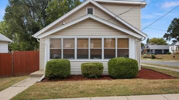 A single-story house with a gabled roof, a front porch, and a small front yard with two bushes.