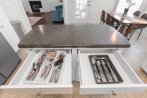 Een keuken met een granieten aanrecht, twee lades met keukengerei, en een eethoek met een houten tafel en stoelen.