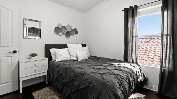 Une chambre à coucher comprenant un lit, une table de chevet, un miroir, un luminaire fixé au mur et une fenêtre avec des rideaux.