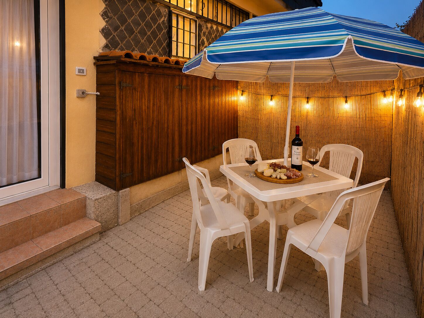 Una terrazza esterna con un tavolo apparecchiato per due, una bottiglia di vino e un piatto di formaggi e salumi.