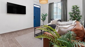 A modern living room with a flat-screen TV mounted on the wall, a blue door, a large window with curtains, a brown leather sofa, and a potted plant. - Apt B, Sunny San Juan Getaway (San Juan)