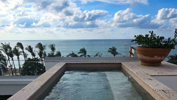 Et rektangulært pool med krystalklart vand, omgivet af en stenterrasse, med udsigt til havet og palmer i baggrunden.