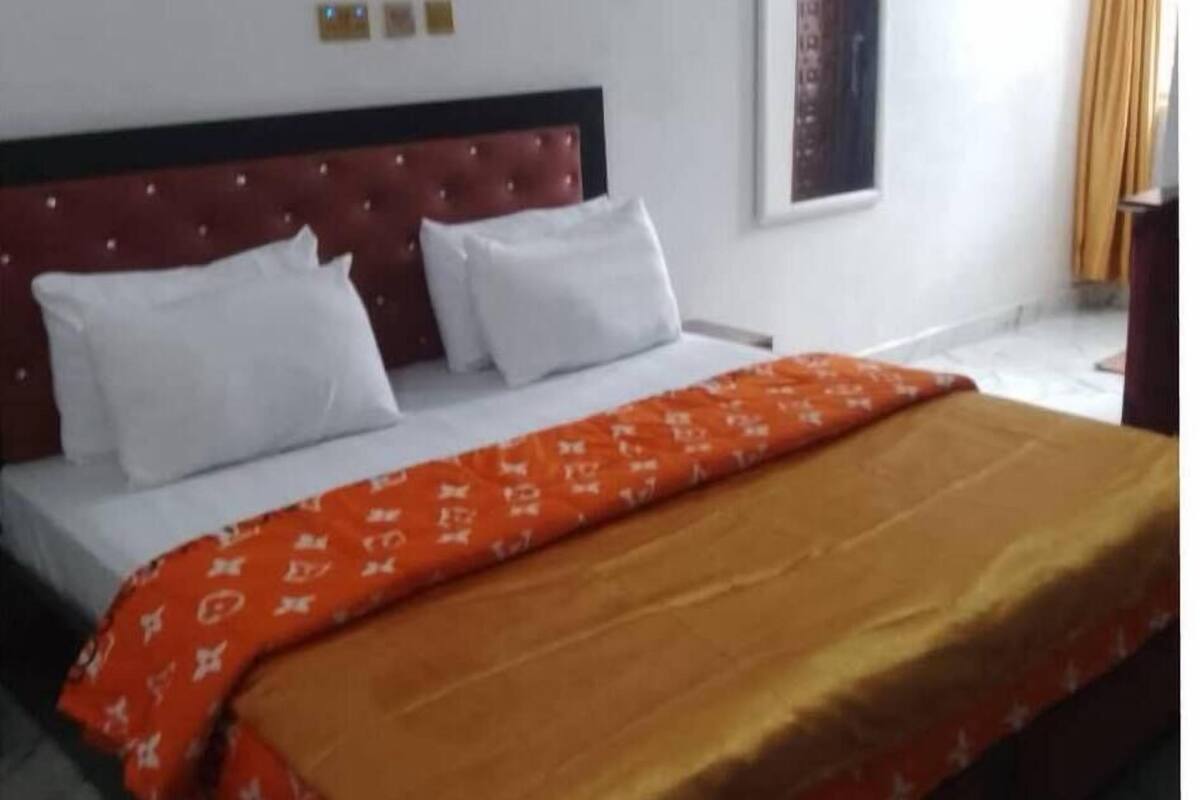 A hotel room with a bed, white pillows, a red headboard, an air conditioner, a window with curtains, and a small table.