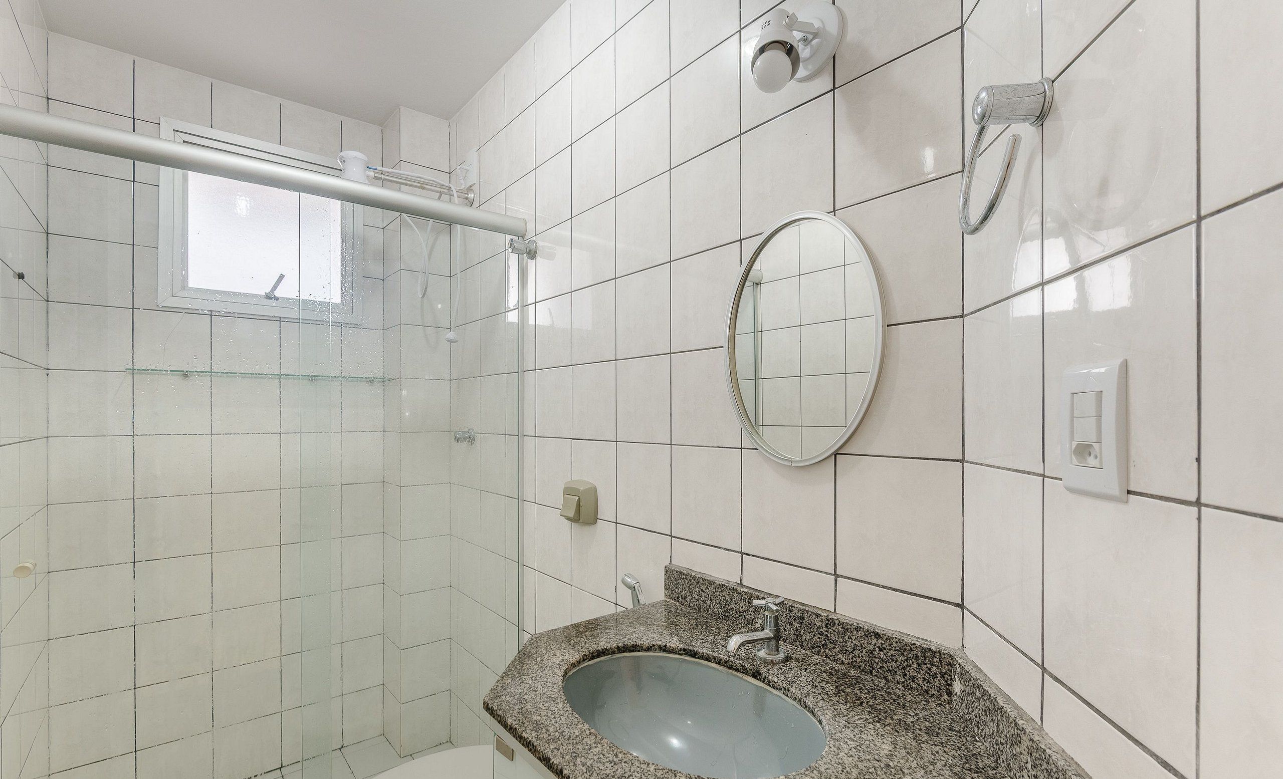 A bathroom with white tiled walls, a granite sink, a mirror, and a shower area.