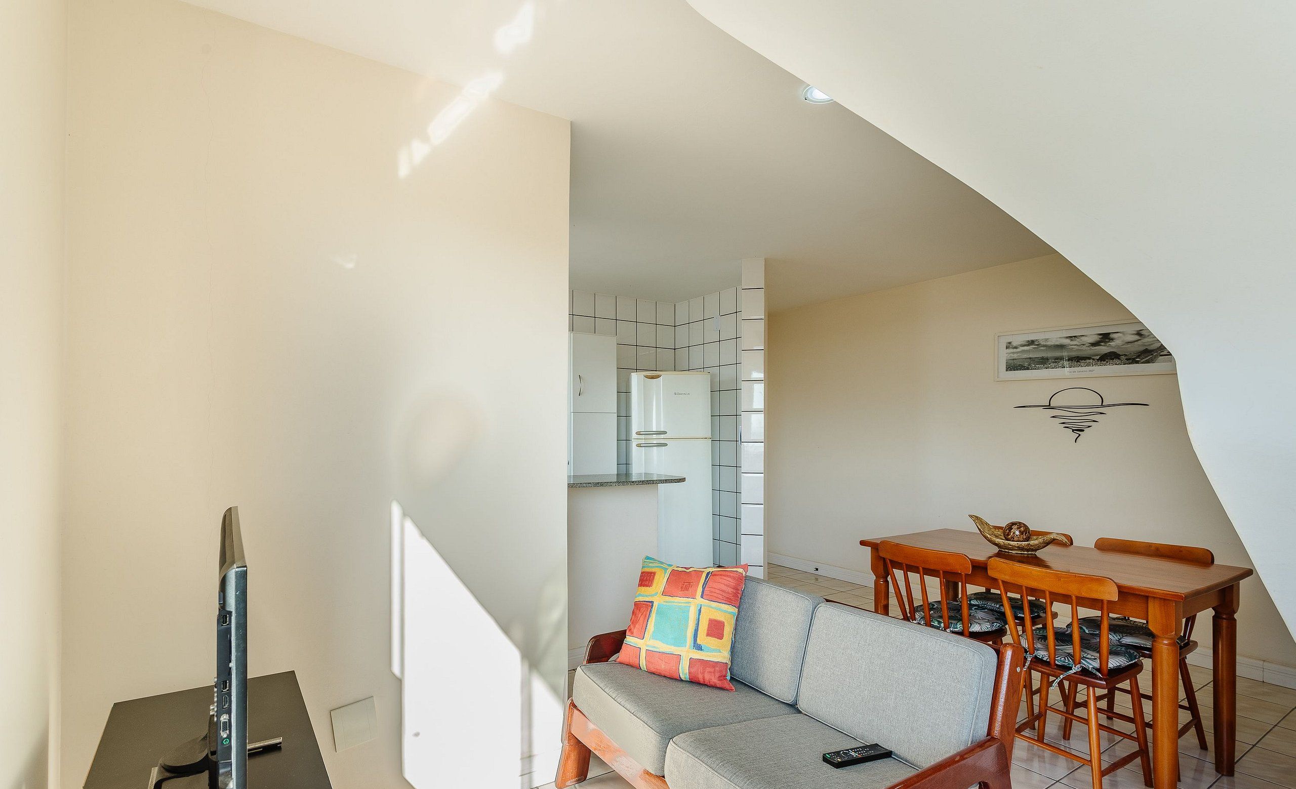 Uma sala de estar moderna com um sofá, uma mesa de jantar de madeira e cadeiras. Uma televisão está posicionada sobre um suporte.