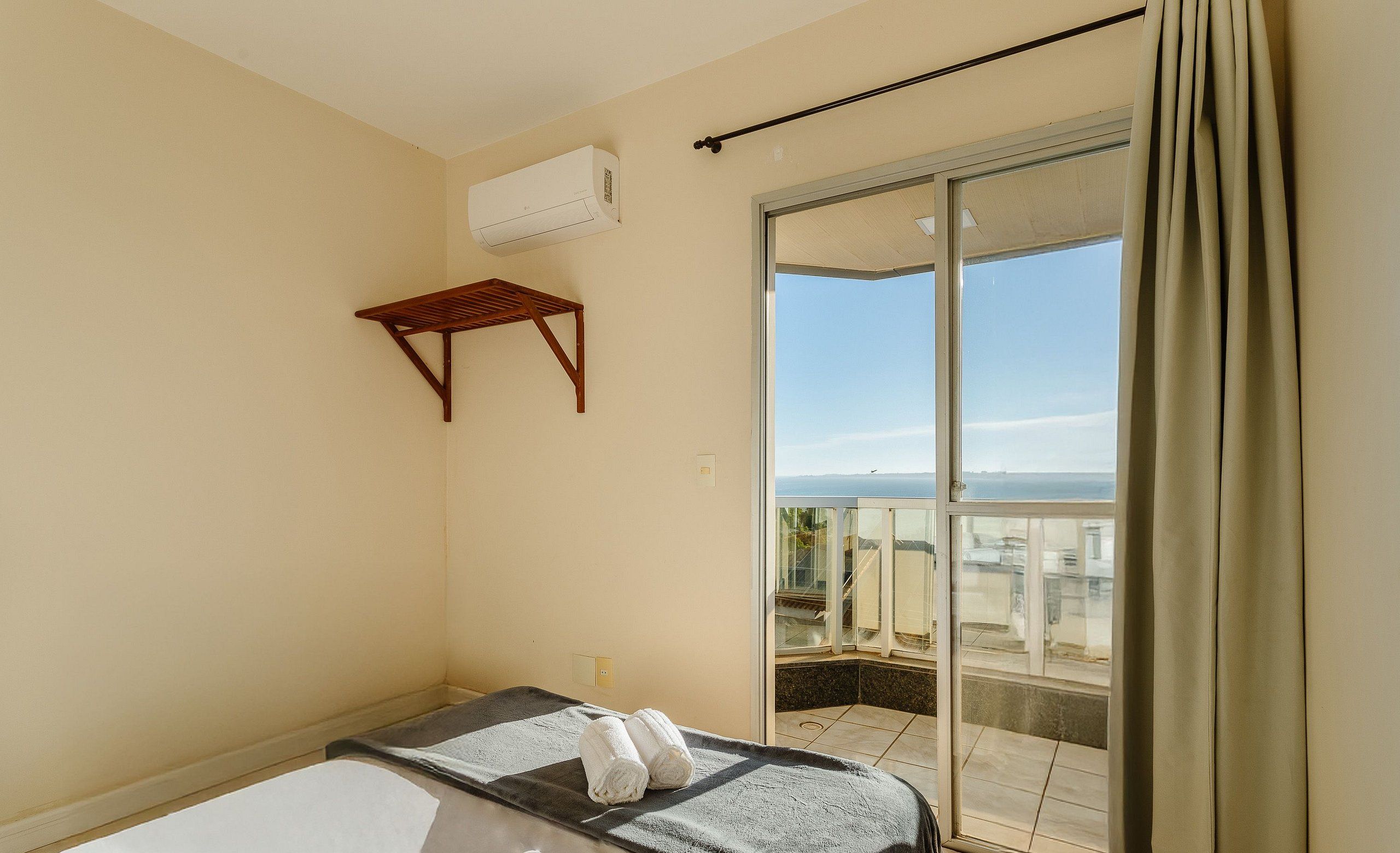 A hotel room with a bed, a small wooden shelf, and a sliding glass door leading to a balcony with a view.
