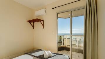 A hotel room with a bed, a small wooden shelf, and a sliding glass door leading to a balcony with a view.