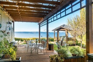 Un patio techado al aire libre con terraza de madera, mobiliario exterior y vistas al agua.