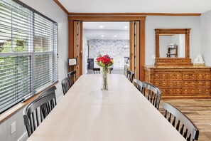 Un comedor con una mesa larga, sillas, un jarrón con flores, un mueble de madera y un espejo.
