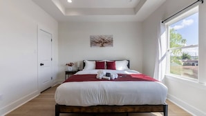 A modern bedroom with a large bed, white walls, a wooden floor, a window with a view, and a door.