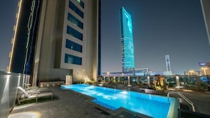 A rooftop pool area with lounge chairs, a swimming pool, and a view of a tall building with illuminated windows. - DAMAC Super Luxury Two Bedroom Apartment (Riyadh)