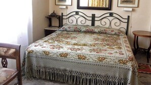 A bedroom with a floral bedspread, a wooden headboard, a small bedside table, a chair, and a mirror on the wall.