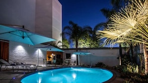 Una piscina con sillones de descanso y sombrillas, rodeada de palmeras y un edificio con fachada blanca.