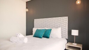 Una cama bien hecha con sábanas blancas y almohadones azul verdosos, una mesita de noche con lámpara y un cabecero estampado.