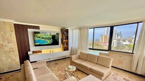 Een woonkamer met een grote flatscreen-tv, een beige bank, een salontafel en een stadszicht door grote ramen.