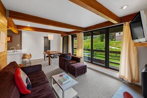 A spacious living area with a sofa, ottoman, and coffee table, a dining table with chairs, and a large window offering a view of greenery. - Base Camp Condo (Crested Butte)