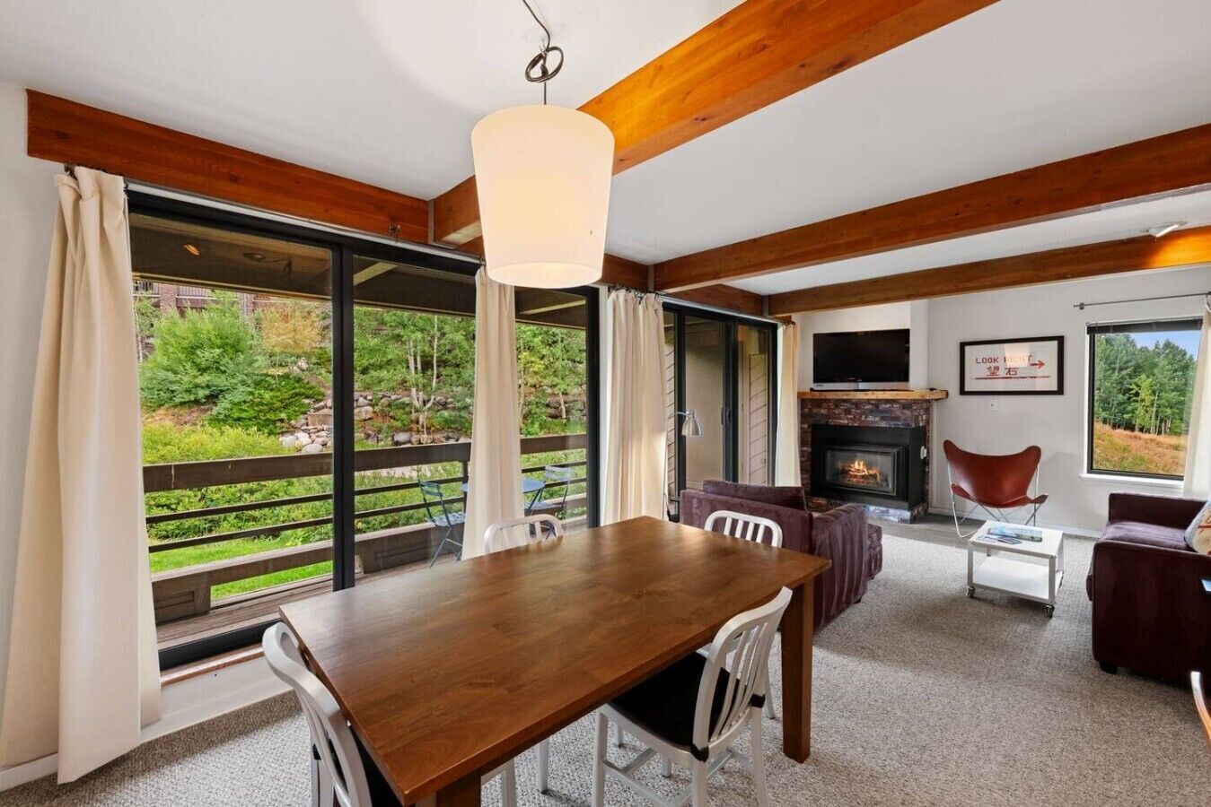 A spacious room with a wooden dining table, a fireplace, and large windows offering a view of a garden.