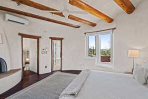 Un dormitorio con una cama grande, vigas de madera, un ventilador de techo, una ventana con vistas y una lámpara de noche.