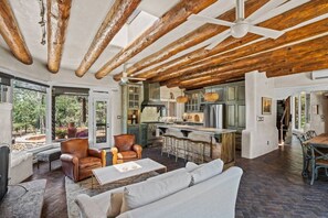A spacious living area with a high ceiling, wooden beams, a large window, and a kitchen with a bar.