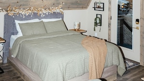 Une chambre avec un plafond en pente, un lit recouvert d’une couette grise et beige, un plancher en bois et une porte donnant sur une voiture garée à l’extérieur.
