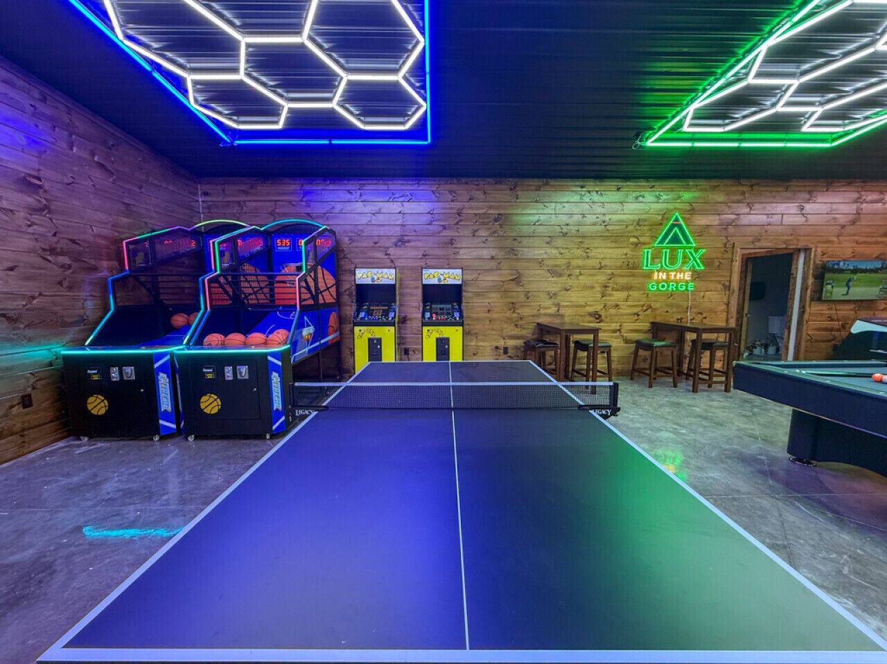 A billiards room with a pool table, neon hexagonal ceiling lights, and arcade machines.