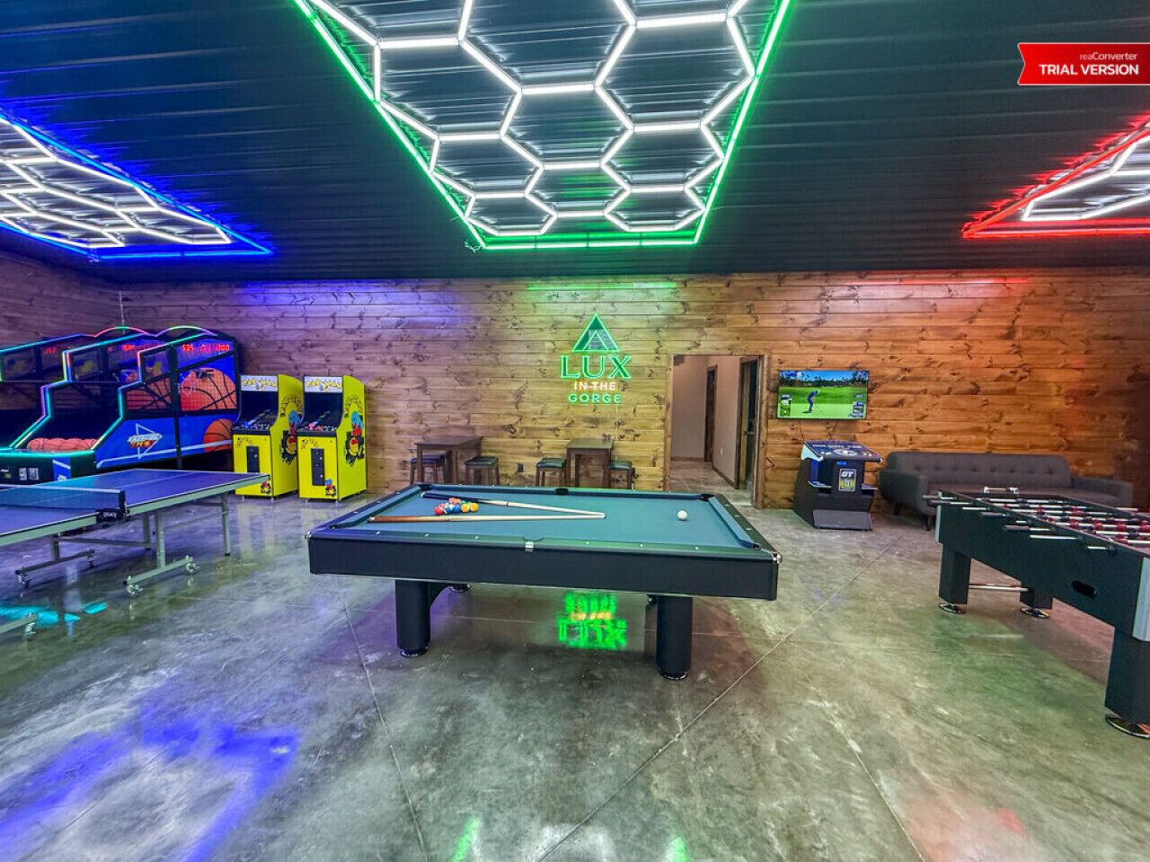 A pool table in the center of a room with neon lights, a foosball table, and arcade games.