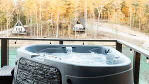 Un jacuzzi en un balcón con vistas a un bosque y dos cabañas al fondo.