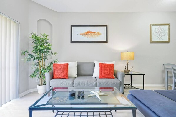 A living room with a grey sofa, red and white cushions, a glass coffee table, a potted plant, and framed artwork on the wall.