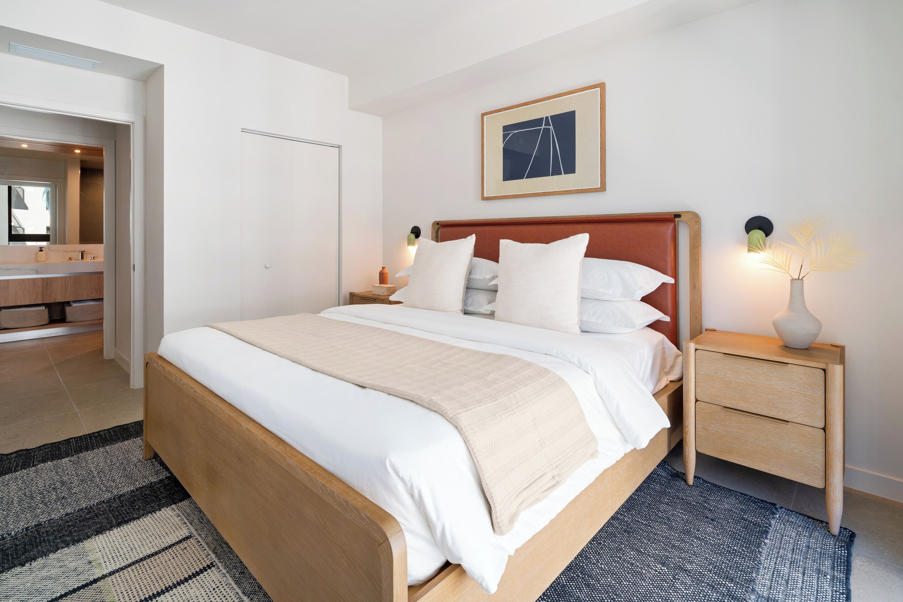 A modern hotel room with a large bed, a bedside table with a lamp, a framed picture on the wall, and a bathroom visible through an open door.