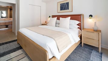A modern hotel room with a large bed, a bedside table with a lamp, a framed picture on the wall, and a bathroom visible through an open door.