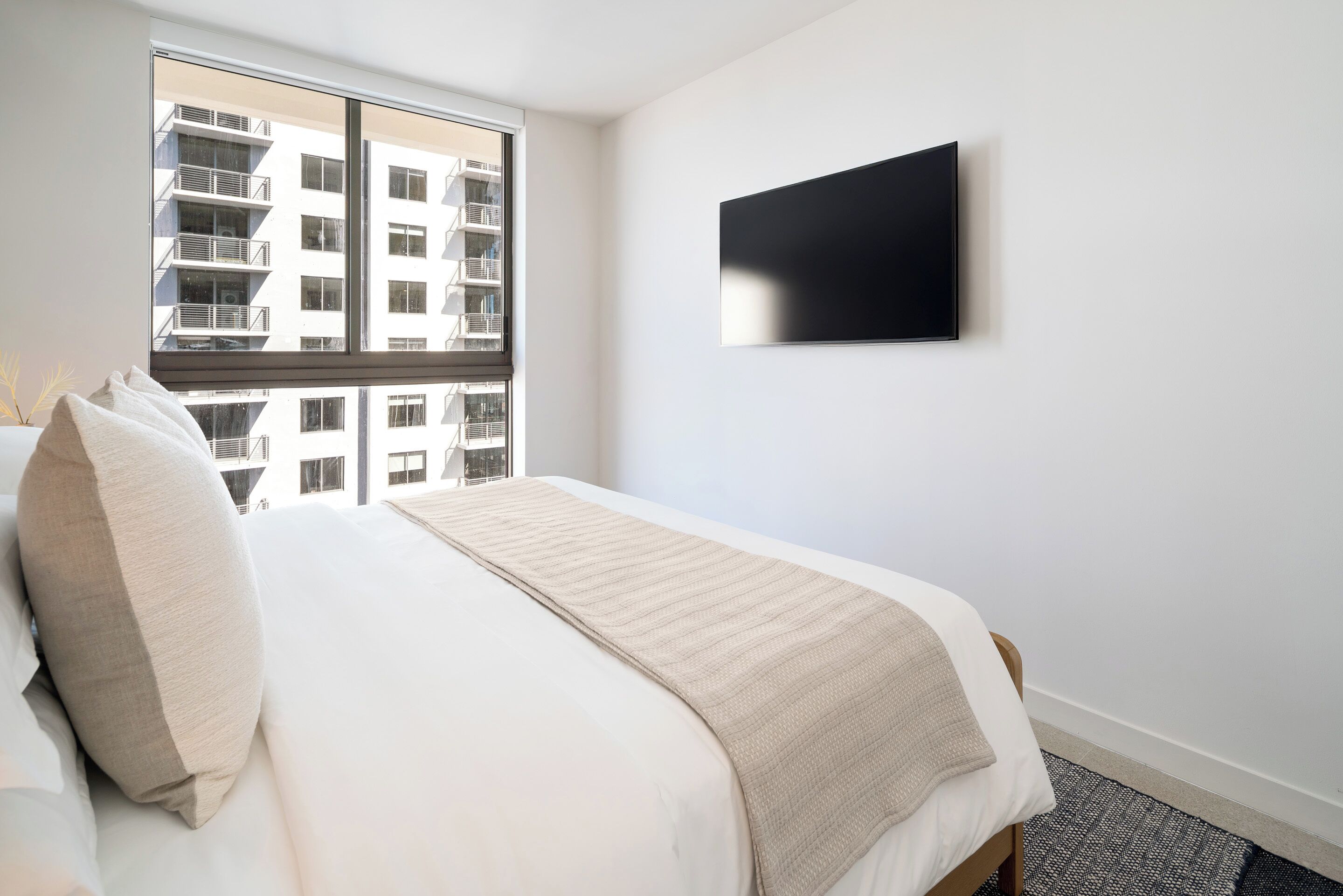 A hotel room with a large bed, a window with a view of a building, and a flat-screen TV mounted on the wall.