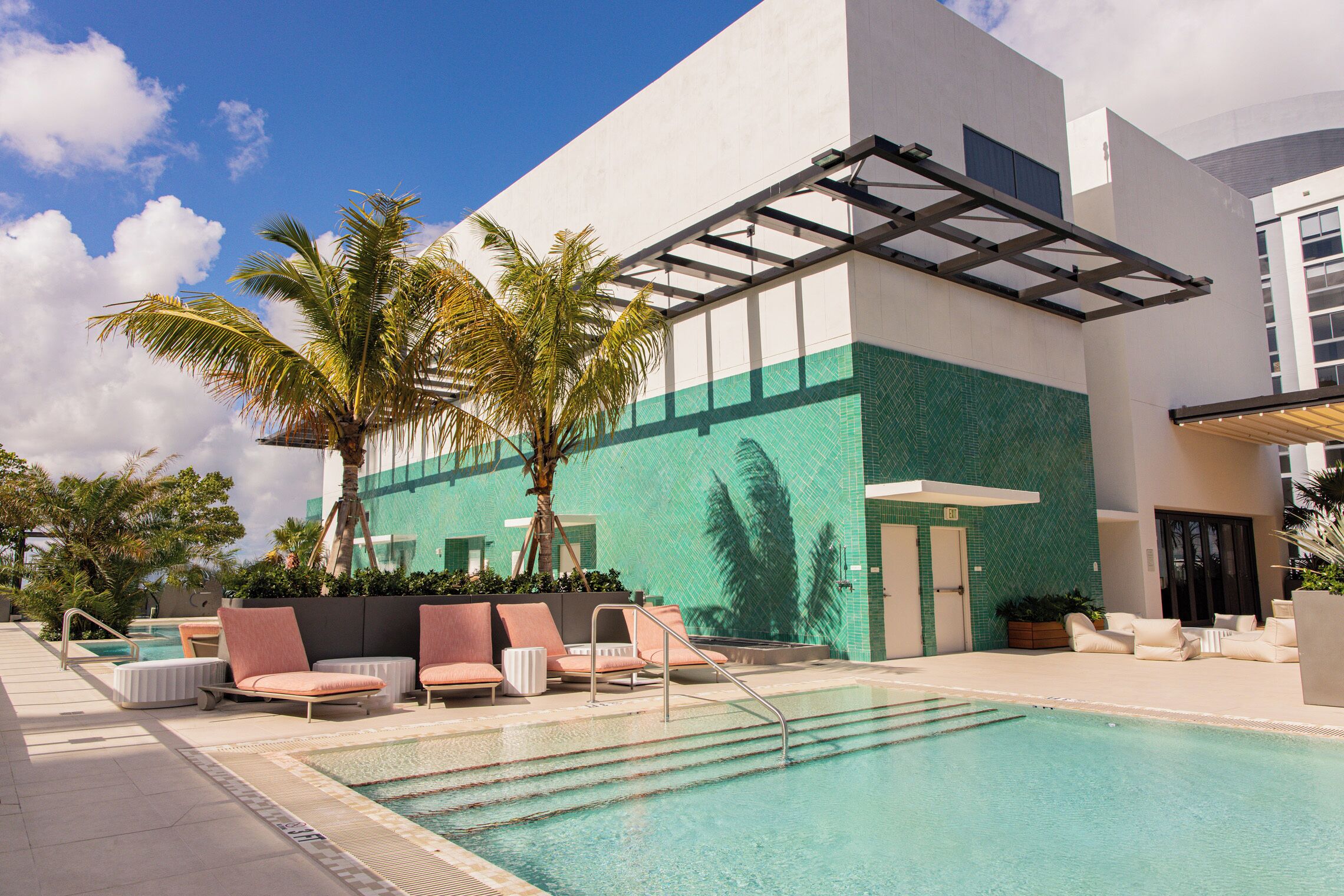 Un espace piscine avec des chaises longues et des palmiers, un coin salon abrité et un bâtiment dont la façade est recouverte de carreaux verts.
