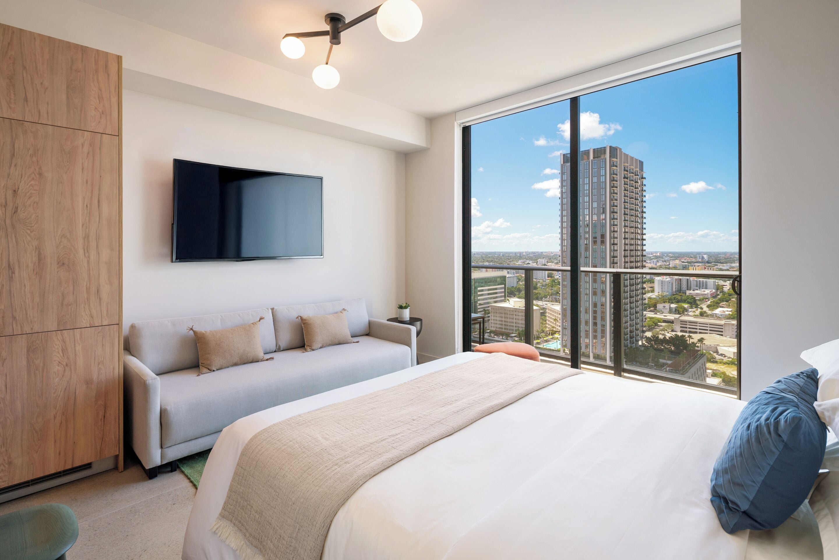 Ein modernes Hotelzimmer mit einem großen Bett, einer Sitzecke, einem an der Wand befestigten Fernseher und einem großen Fenster mit Blick auf die Stadt.