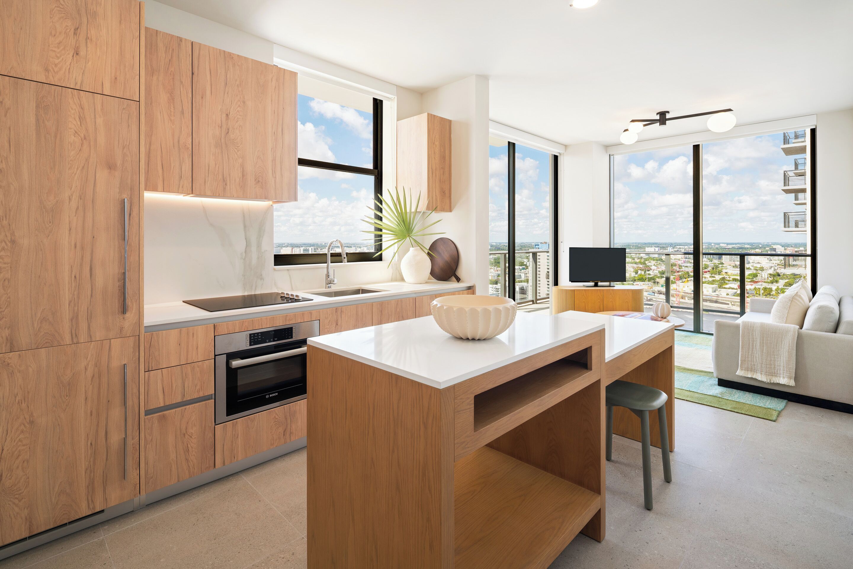 A modern kitchen with wooden cabinets, a central island, and a large window offering a city view.