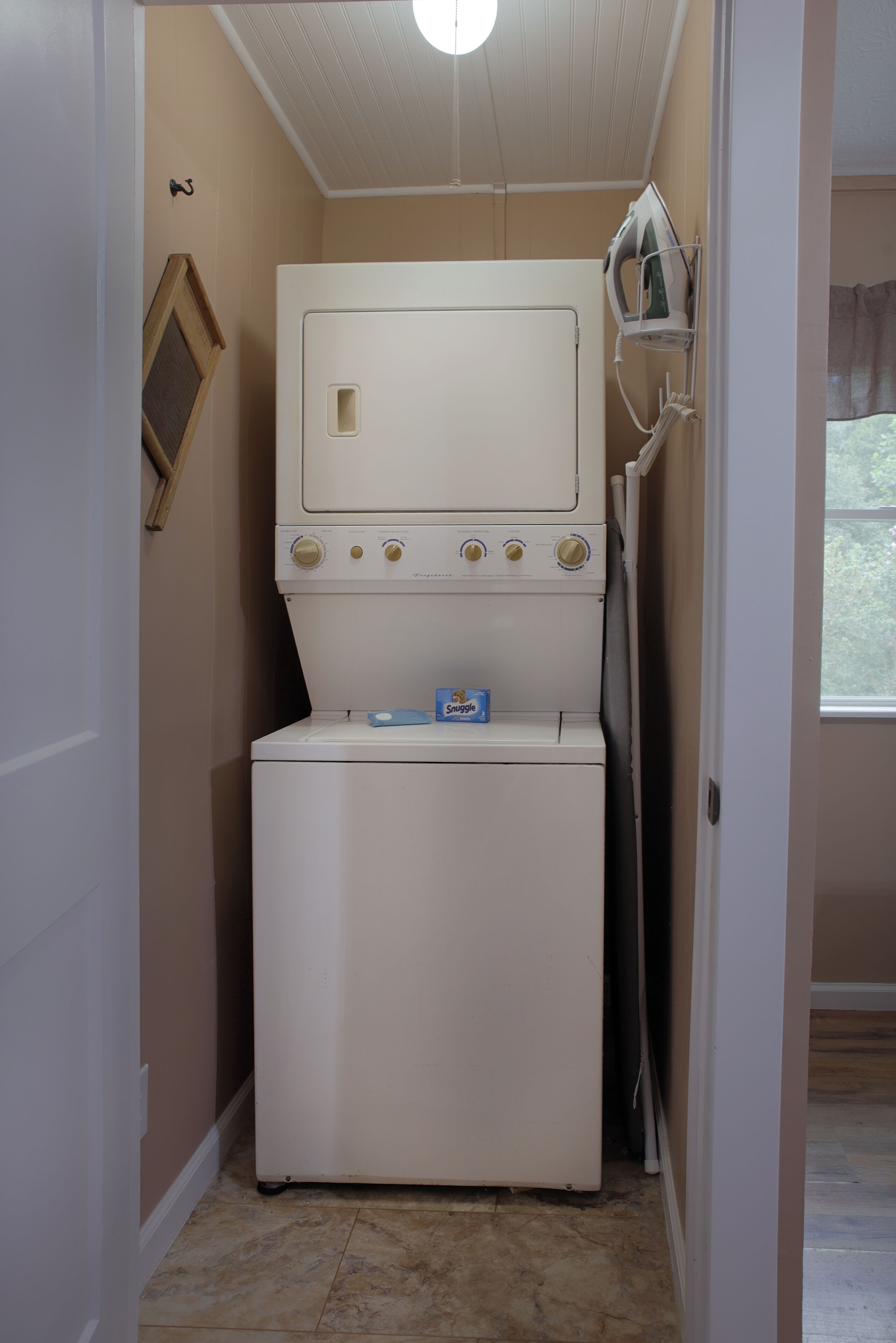 A compact laundry room with a stacked washer and dryer, a small ironing board, and a framed picture on the wall.