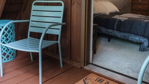 Interior acogedor de una cabaña con techo de madera, una cama con manta azul oscuro, una silla metálica azul claro y una puerta de madera.