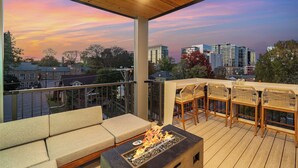 Un área de estar al aire libre techada con chimenea, asientos acolchados y vista a un paisaje urbano al atardecer.